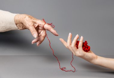 Hands of a young and an senior woman reach out to each other, connected by a red thread